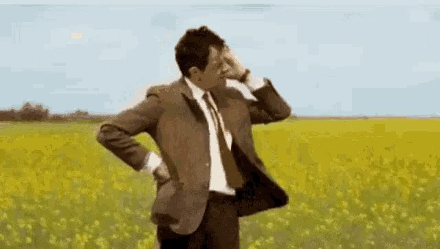 Un uomo in giacca e cravatta in un campo di fiori gialli si mette una mano sulla fronte, in attesa